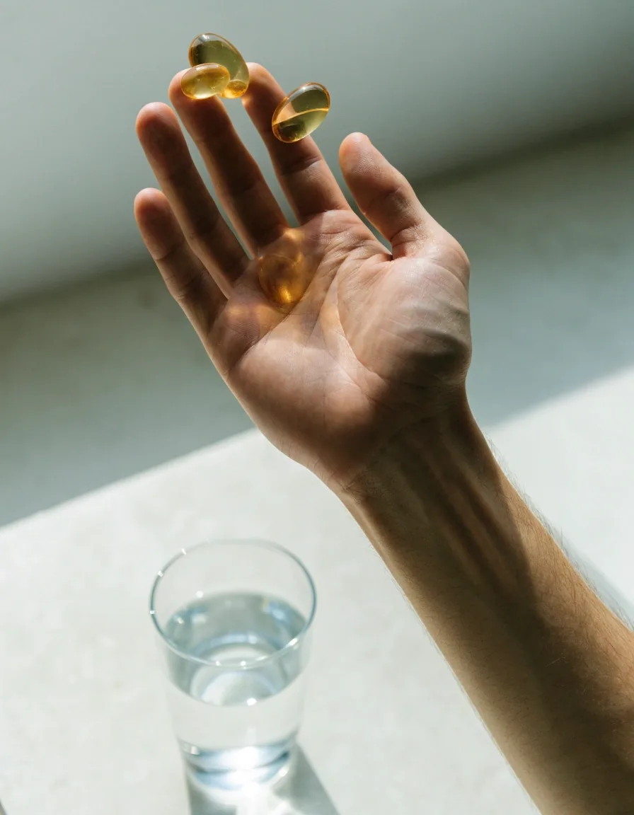 Hands beside a laptop with a faint page glow, a glass of water and a small dish of capsules on a pale counter
